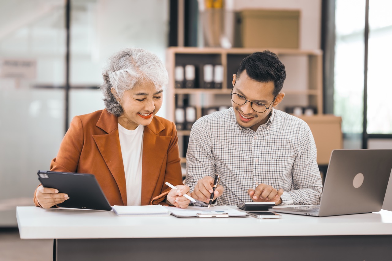 An older mature woman talks to asian man. They have a laptop and papers. The man looks stressed.